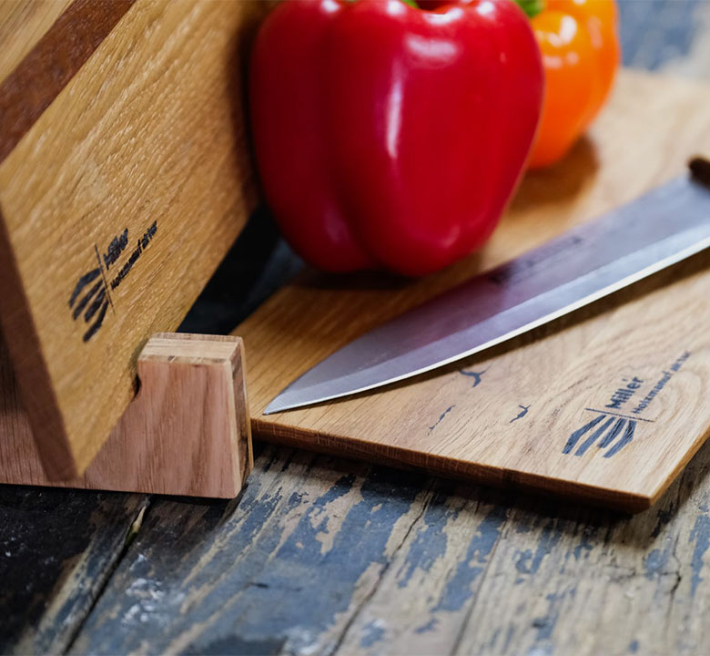 Ein Holz-Messerblock und zwei Schneidebretter aus Holz mit einem Messer darauf, daneben eine rote und eine orange Paprika auf einer rustikalen Holzoberfläche.